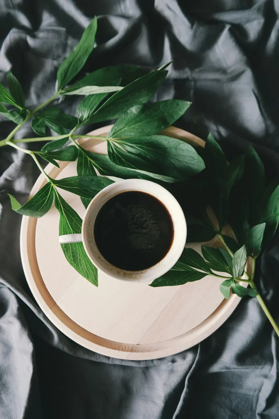 A small white cup of black coffee on a wooden tray with peony leaves on grey linen.