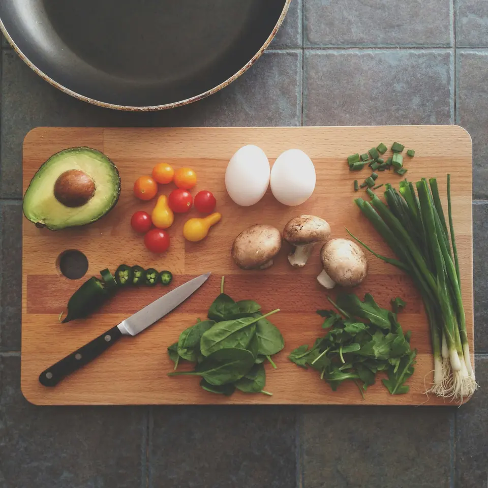 A wooden chopping board with avocado, eggs, mushrooms, spinach, tomatoes and spring onions.