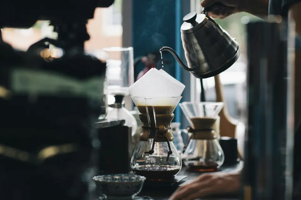 A pour-over Chemex setup with a gooseneck kettle pouring water onto coffee grounds.